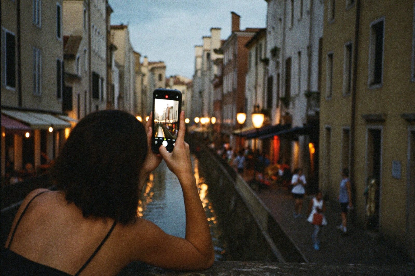 A traveler photographing a lantern-lit canal at blue hour, the shot framed on her glowing phone