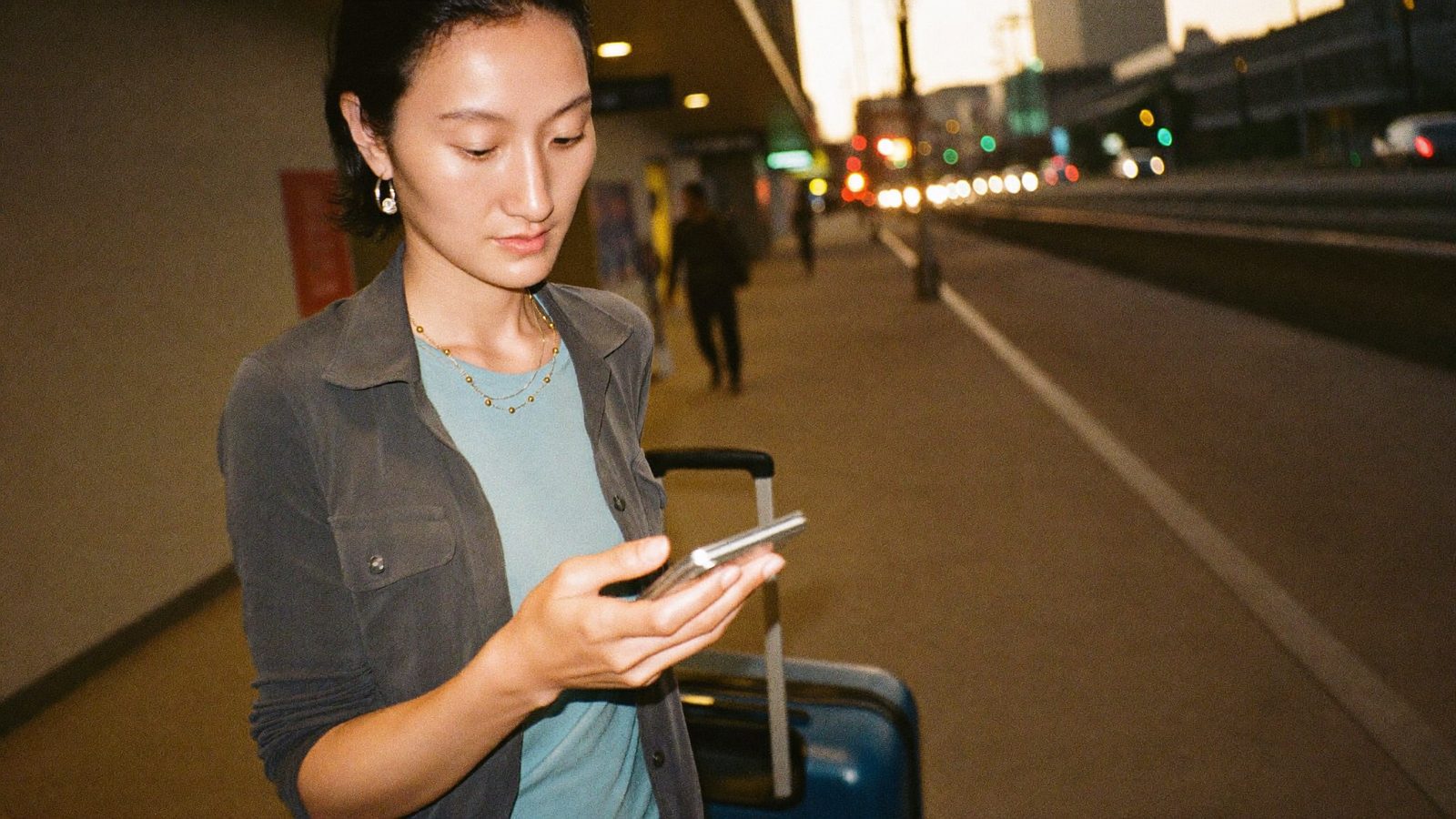 A traveler outside the arrivals hall at night, checking her phone beside her suitcase - already connected